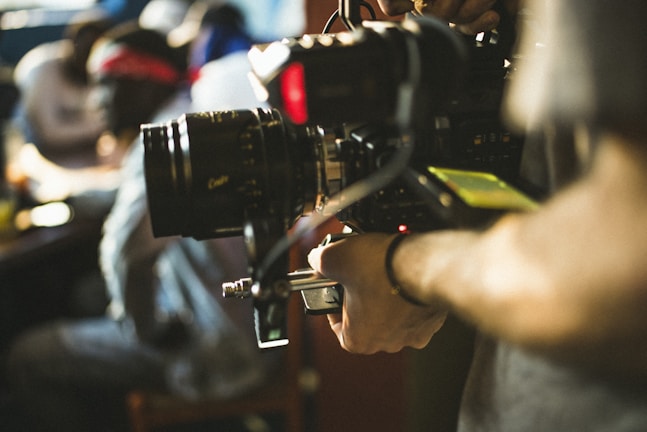 A person operating a professional video camera, with a blurred background suggesting an indoor setting. The camera appears large and complex, indicating use in filmmaking or broadcasting.