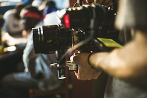 A person operating a professional video camera, with a blurred background suggesting an indoor setting. The camera appears large and complex, indicating use in filmmaking or broadcasting.