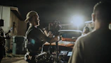 A dynamic behind-the-scenes shot of a film crew setting up lights on a rustic outdoor set at dusk.