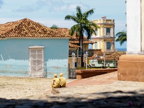 A cobblestone street extends towards colorful colonial-style buildings, featuring a blue wall with a white door and washed-out paint. Two palm trees rise prominently in the background, amid other vibrant structures decorated in rustic yellows and blues. In the foreground, two yellow sculptures of people sit, seemingly watching the scenery.