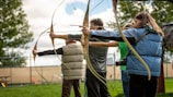 Several people are lined up outdoors, holding bows and arrows, aiming at a target. They are dressed in casual clothing with one person wearing a blue vest and another in a light-brown padded vest. There is greenery and trees in the background along with a chain-link fence and some outdoor seating.