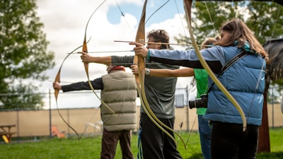 Several people are lined up outdoors, holding bows and arrows, aiming at a target. They are dressed in casual clothing with one person wearing a blue vest and another in a light-brown padded vest. There is greenery and trees in the background along with a chain-link fence and some outdoor seating.