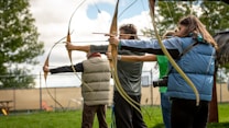 Several people are lined up outdoors, holding bows and arrows, aiming at a target. They are dressed in casual clothing with one person wearing a blue vest and another in a light-brown padded vest. There is greenery and trees in the background along with a chain-link fence and some outdoor seating.