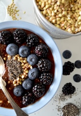 A bowl of breakfast porridge topped with blackberries, blueberries, buckwheat, and chia seeds. The spoon rests inside the bowl, and a separate container of buckwheat is nearby, along with some scattered seeds and berries on the surface.