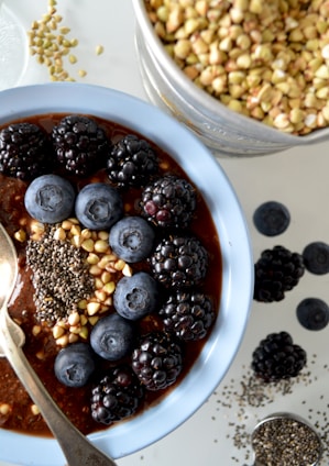 A bowl of breakfast porridge topped with blackberries, blueberries, buckwheat, and chia seeds. The spoon rests inside the bowl, and a separate container of buckwheat is nearby, along with some scattered seeds and berries on the surface.