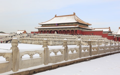 A large historical palace complex with traditional Chinese architecture, featuring red walls, ornate golden roofs, and decorative stone railings. The ground is covered in snow, and numerous people are walking along the pathways.