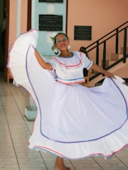 A woman is engaged in traditional dance, wearing a flowing white dress with red and blue trim. Her hair is adorned with flowers, and she is accessorized with large circular earrings. Her eyes are closed and she appears to be in mid-movement, with her dress spread wide, suggesting a spinning motion. She stands inside a building, with plaques on the wall and stairs in the background.