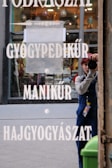 A person wearing dark clothing and a cap is taking a photo of their reflection in a glass window. The window displays white text with words like 'GYÓGYPEDIKŰR,' 'MANIKŰR,' and 'HAJGYÓGYÁSZAT.' Inside, hanging lamps and other blurred elements are visible. A green trash bin is partially visible on the lower right.