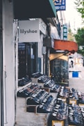 A storefront display featuring rows of shoes arranged on wooden shelves outside a shop called lilyshoe. The store is located on a street with other neighboring shops visible and a sidewalk along the side.