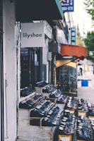 A storefront display featuring rows of shoes arranged on wooden shelves outside a shop called lilyshoe. The store is located on a street with other neighboring shops visible and a sidewalk along the side.