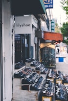 A storefront display featuring rows of shoes arranged on wooden shelves outside a shop called lilyshoe. The store is located on a street with other neighboring shops visible and a sidewalk along the side.