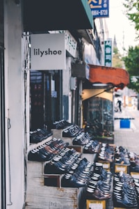 A storefront display featuring rows of shoes arranged on wooden shelves outside a shop called lilyshoe. The store is located on a street with other neighboring shops visible and a sidewalk along the side.