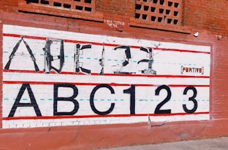 A mural on a brick wall features an alphabet and numbers, with the upper line formed by weapons shaped to resemble letters. Below, the large, bold text 'ABC123' is painted in black. Red and blue lines run horizontally across the mural, giving it the appearance of a lined notebook sheet. The backdrop is a red brick wall with some decorative brickwork at the top. The word 'FURTIVE' appears on the mural as part of the art.