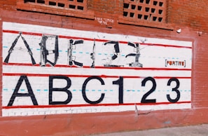 A mural on a brick wall features an alphabet and numbers, with the upper line formed by weapons shaped to resemble letters. Below, the large, bold text 'ABC123' is painted in black. Red and blue lines run horizontally across the mural, giving it the appearance of a lined notebook sheet. The backdrop is a red brick wall with some decorative brickwork at the top. The word 'FURTIVE' appears on the mural as part of the art.