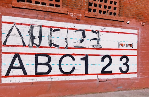 A mural on a brick wall features an alphabet and numbers, with the upper line formed by weapons shaped to resemble letters. Below, the large, bold text 'ABC123' is painted in black. Red and blue lines run horizontally across the mural, giving it the appearance of a lined notebook sheet. The backdrop is a red brick wall with some decorative brickwork at the top. The word 'FURTIVE' appears on the mural as part of the art.