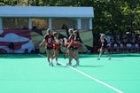 A group of female lacrosse players in black and orange uniforms are gathered together on a green field, likely discussing strategy or celebrating. In the background, additional team members are on the sidelines near a structure resembling a tent or pavilion. Trees are visible beyond the pavilion, suggesting an outdoor setting.