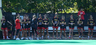 A group of female athletes in matching sports uniforms are standing together, some holding lacrosse sticks. They appear to be part of a team and are positioned on the sidelines of a playing field. The setting is outdoors with trees and a fence in the background. The team members are wearing matching jerseys with the name 'Maryland' and numbers on them, and some are wearing glasses or protective eyewear.