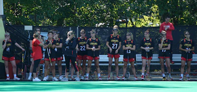 A group of female athletes in matching sports uniforms are standing together, some holding lacrosse sticks. They appear to be part of a team and are positioned on the sidelines of a playing field. The setting is outdoors with trees and a fence in the background. The team members are wearing matching jerseys with the name 'Maryland' and numbers on them, and some are wearing glasses or protective eyewear.
