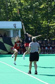 Two athletes are engaged in a dynamic play during a lacrosse game, reaching high with their sticks to catch a ball in mid-air. A referee in a black and white striped shirt stands nearby, overseeing the action. The game takes place on a vibrant green field with trees in the background.