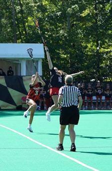 Two athletes are engaged in a dynamic play during a lacrosse game, reaching high with their sticks to catch a ball in mid-air. A referee in a black and white striped shirt stands nearby, overseeing the action. The game takes place on a vibrant green field with trees in the background.