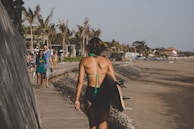 A person in a swimsuit with a surfboard walks along a beachside path. The scene includes palm trees, various beachgoers, and buildings nearby. The sky is clear, and the sun casts a warm glow over the area.