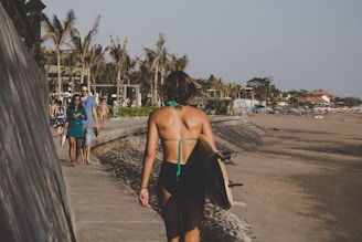 A person in a swimsuit with a surfboard walks along a beachside path. The scene includes palm trees, various beachgoers, and buildings nearby. The sky is clear, and the sun casts a warm glow over the area.