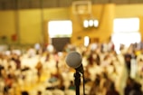 A close-up view of a microphone in focus with a large, blurred audience in the background. The setting appears to be an indoor venue, possibly a conference or event, with people seated and light streaming through windows.