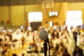 A close-up view of a microphone in focus with a large, blurred audience in the background. The setting appears to be an indoor venue, possibly a conference or event, with people seated and light streaming through windows.