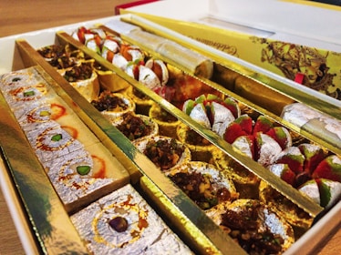 An assortment of colorful Indian sweets neatly arranged in a decorative box. The sweets are various shapes and sizes, some adorned with silver leaf and nuts. The box has multiple compartments to separate the different types of confections.