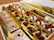 An assortment of colorful Indian sweets neatly arranged in a decorative box. The sweets are various shapes and sizes, some adorned with silver leaf and nuts. The box has multiple compartments to separate the different types of confections.