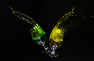 Two glasses with colored liquid in them, one with green liquid and the other with yellow, are captured in mid-air with the liquid splashing outward against a black background.