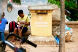 A man wearing a yellow shirt is seated by a large, weathered mailbox that has text in multiple languages including French and an Asian language. He is holding a piece of fruit and appears to be peeling it. Beside him is a red and black backpack, and a young boy in a blue shirt stands nearby facing away. The background shows a tree with large green leaves and a river, while the foreground includes a bicycle and a basket of plastic bottles and trash.