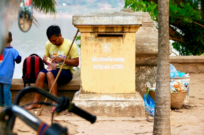 A man wearing a yellow shirt is seated by a large, weathered mailbox that has text in multiple languages including French and an Asian language. He is holding a piece of fruit and appears to be peeling it. Beside him is a red and black backpack, and a young boy in a blue shirt stands nearby facing away. The background shows a tree with large green leaves and a river, while the foreground includes a bicycle and a basket of plastic bottles and trash.
