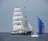 A large sailing ship with tall white sails bearing the phrase 'WIND IS OUR FRIEND' navigates the ocean alongside a smaller sailboat with a bright blue spinnaker sail. The sea is calm and the sky is clear, creating a serene maritime scene.