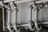 A series of interconnected industrial pipes with visible joints and flanges. The pipes have a worn, matte texture and are arranged in a horizontally aligned, organized manner. The background is an industrial setting, possibly a basement or maintenance area, with a neutral color palette.
