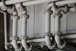 A series of interconnected industrial pipes with visible joints and flanges. The pipes have a worn, matte texture and are arranged in a horizontally aligned, organized manner. The background is an industrial setting, possibly a basement or maintenance area, with a neutral color palette.