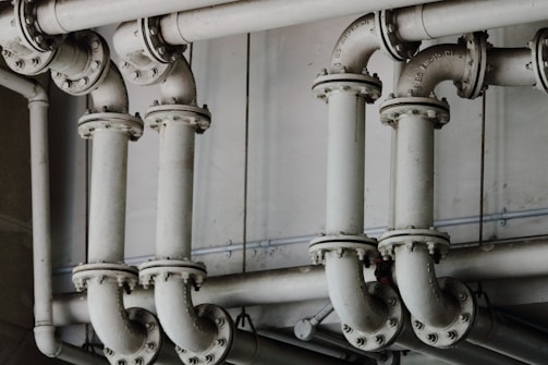 A series of interconnected industrial pipes with visible joints and flanges. The pipes have a worn, matte texture and are arranged in a horizontally aligned, organized manner. The background is an industrial setting, possibly a basement or maintenance area, with a neutral color palette.