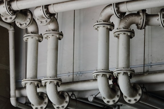 A series of interconnected industrial pipes with visible joints and flanges. The pipes have a worn, matte texture and are arranged in a horizontally aligned, organized manner. The background is an industrial setting, possibly a basement or maintenance area, with a neutral color palette.