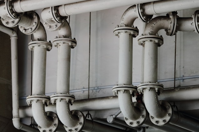 A series of interconnected industrial pipes with visible joints and flanges. The pipes have a worn, matte texture and are arranged in a horizontally aligned, organized manner. The background is an industrial setting, possibly a basement or maintenance area, with a neutral color palette.