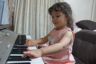 A young child with curly hair is sitting and playing an electronic keyboard. The child is wearing a pink dress with a bow detail and seems focused on the keys. The setting appears to be indoors with curtains in the background and part of a couch visible.