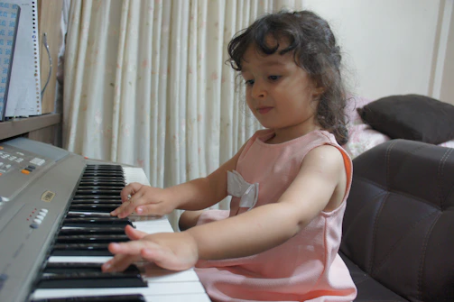 A young child with curly hair is sitting and playing an electronic keyboard. The child is wearing a pink dress with a bow detail and seems focused on the keys. The setting appears to be indoors with curtains in the background and part of a couch visible.