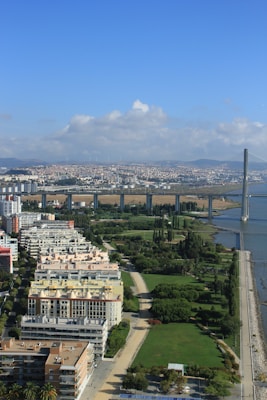Aerial view of an urban area with high-rise apartment buildings, well-maintained green spaces with trees, and a prominent bridge spanning across a river. The cityscape continues in the background with scattered clouds under a clear blue sky.