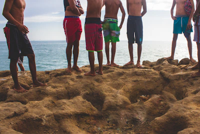 A group of friends laughing and wearing Kalima apparel near a rocky shoreline.