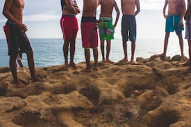 A group of shirtless individuals standing on rugged, rocky terrain near a body of water. They are wearing colorful swim trunks and are enjoying the beach setting, with the ocean visible in the background.