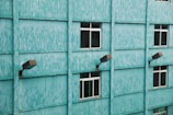A teal-colored building facade with a textured pattern, featuring several uniformly spaced windows and outdoor lighting fixtures attached to the wall.