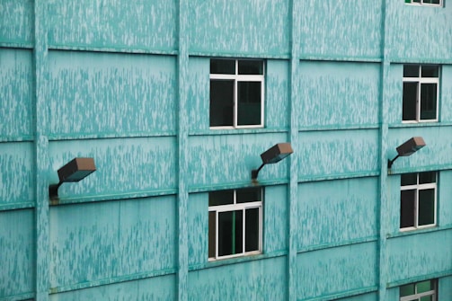 A teal-colored building facade with a textured pattern, featuring several uniformly spaced windows and outdoor lighting fixtures attached to the wall.