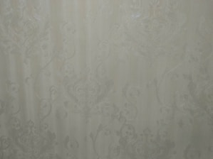 A close-up view of an intricate wallpaper pattern featuring ornate, swirling designs with floral and leaf motifs in a subtle, embossed texture. The background is a soft, muted beige color, giving it an elegant and classic appearance.