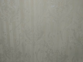 A close-up view of an intricate wallpaper pattern featuring ornate, swirling designs with floral and leaf motifs in a subtle, embossed texture. The background is a soft, muted beige color, giving it an elegant and classic appearance.