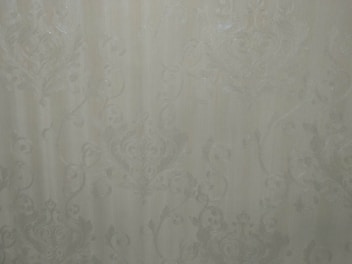 A close-up view of an intricate wallpaper pattern featuring ornate, swirling designs with floral and leaf motifs in a subtle, embossed texture. The background is a soft, muted beige color, giving it an elegant and classic appearance.