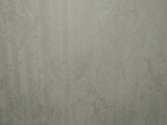 A close-up view of an intricate wallpaper pattern featuring ornate, swirling designs with floral and leaf motifs in a subtle, embossed texture. The background is a soft, muted beige color, giving it an elegant and classic appearance.